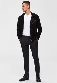 Black suede biker jacket with zippered pockets, white V-neck t-shirt, and dark trousers. He wears black lace-up shoes.