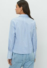 Chemise rayée bleu clair avec un col ajusté, des manches longues, des poignets boutonnés et un ourlet arrondi. Le tissu semble lisse et léger.