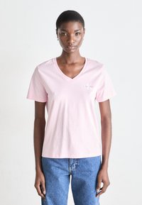 Rožinė medvilninė t-shirt su V formos iškirpte. Su trumpomis rankovėmis ir mažu baltu logotipu kairėje krūtinės pusėje. Suderinta su mėlynomis džinsinėmis kelnėmis.