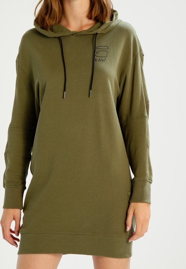 Femme portant une robe sweat à capuche ample de couleur vert olive avec manches longues et cordons noirs.