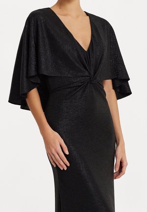 Kobieta w czarnej teksturowanej sukience z głębokim dekoltem w serek i szerokimi, drapowanymi rękawami pelerynowymi, z dekoracyjnym skręconym węzłem w talii.