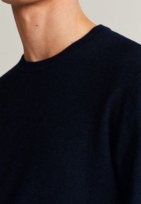 Detailní záběr osoby se sukní s kulatým výstřihem v černé barvě, zachycující krk a horní část hrudníku na neutrálním pozadí.