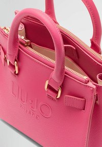 Sac à main synthétique rose avec surface texturée, matériel doré, doubles poignées et fermeture zippée. Présente un logo embossé sur le devant.