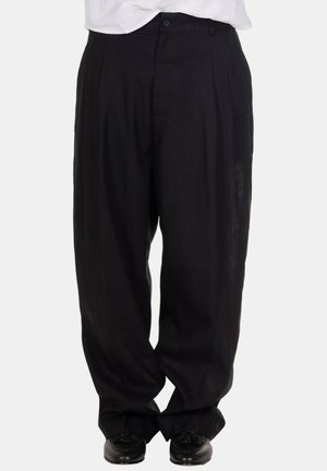 Uomo con pantaloni neri larghi plissettati, mocassini neri con nappine e camicia bianca infilata, in piedi davanti a uno sfondo semplice.
