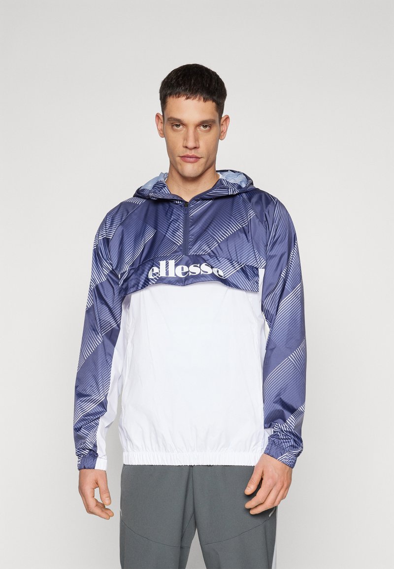 Veste coupe-vent bleu marine et blanche avec demi-fermeture éclair, manches à motifs, poignets élastiques et logo sur la poitrine. Matériau léger, coupe décontractée.