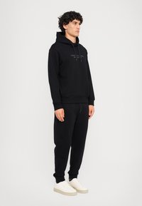 Schwarzer Kapuzenpullover mit Kordelzug, Kängurutasche und Logo auf der Brust; kombiniert mit passenden schwarzen Jogginghosen und weißen Sneakers. Material aus Baumwollmischung.