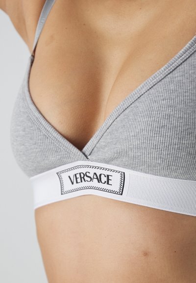 Grå ribbet bralette med V-hals design, med et hvitt elastisk bånd merket "VERSACE." Stoffet har et mykt, teksturert utseende.