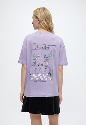 Ξανθιά γυναίκα που φοράει ένα μωβ μπλουζάκι με πολύχρωμη εικονογράφηση χυμού και μια μαύρη φούστα, στέκεται με την πλάτη στην κάμερα.