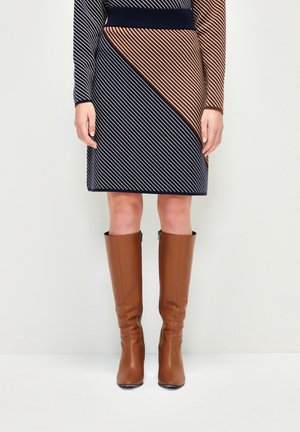 Femme portant une robe à rayures diagonales bleu marine et marron et des bottes en cuir marron montant jusqu'aux genoux, debout contre un mur blanc.