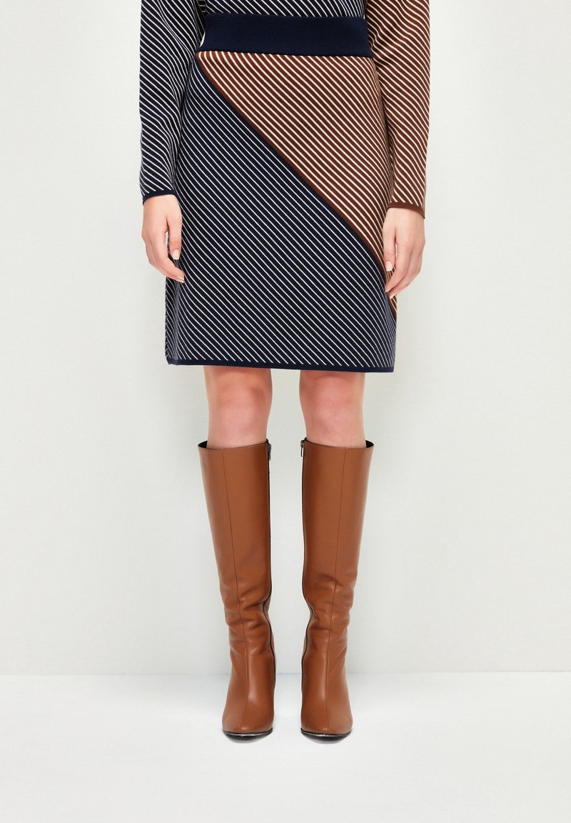 Femme portant une robe à rayures diagonales bleu marine et marron et des bottes en cuir marron montant jusqu'aux genoux, debout contre un mur blanc.