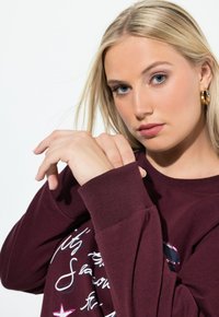 Burgundowa bluza z długim rękawem, z prążkowanym mankietem, grafiką tekstową i akcentem w postaci cekinów na przodzie. Widoczna miękka faktura materiału.