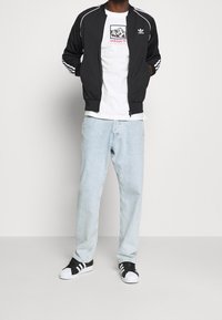 Black zippered jacket with white stripes, white graphic t-shirt, light blue denim jeans, and black and white sneakers with three stripes.