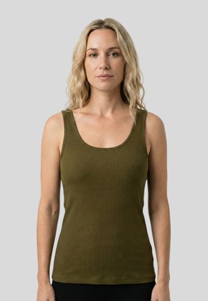 Blonde Frau mit welligem Haar, die ein olivgrünes geripptes ärmelloses Oberteil trägt, steht vor einem schlichten hellgrauen Hintergrund.