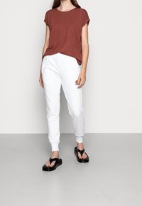 Kvinna iförd en rostfärgad kortärmad topp, vita joggerbyxor med en logotyp och svarta platåsandaler mot en enkel grå bakgrund.