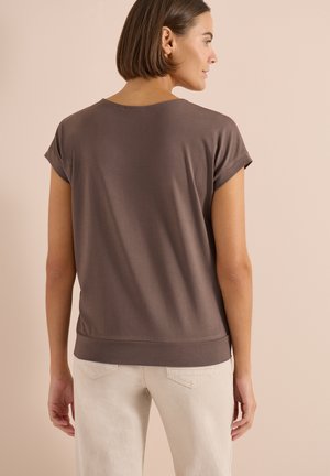 Femme aux cheveux courts bruns portant un haut marron à manches courtes et un pantalon beige clair, debout devant un fond beige uni.
