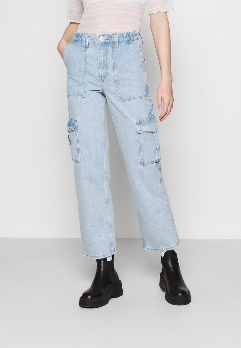 Osoba v světle modrých vysokých cargo džínách s velkými kapsami, průsvitném bílém topu a černých robustních kotníkových botách.