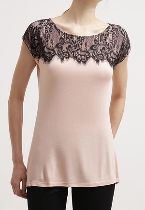 Vrouw draagt een lichtroze mouwloze top met zwart bloemmotief kantdetail over de schouders en de bovenkant van de borst.
