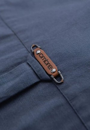 Tissu bleu foncé avec une texture semblable au lin. Présente une étiquette en cuir marron étiquetée "ombre" et des accessoires en argent.