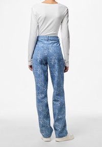 Person som bär en vit långärmad topp och högmidjade blå jeans med ett ros-mönster, sedd från ryggen, står framför en vit bakgrund.