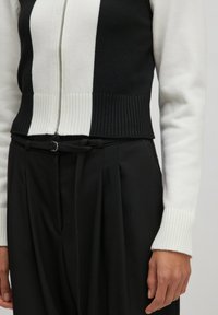 Gebreide cropped zip-up sweater in zwart en wit, met geribde onderkant, lange mouwen en een gladde textuur. Gepaard met hoge zwarte broek.