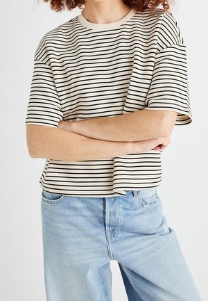 Person mit lockigem Haar, die ein kurzärmeliges, creme-schwarz gestreiftes Hemd und hellblaue Jeans trägt, die Arme vor der Taille verschränkt.