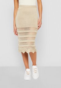Beige Strick-Midirock mit horizontalen Lochmuster und gewelltem Saum. Kombiniert mit weißen Sneakers mit goldenen Akzenten.