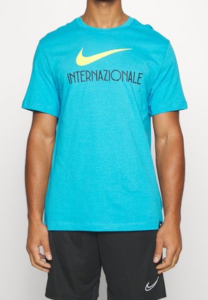 Homme portant un T-shirt turquoise Nike avec le texte "Internazionale" et un short noir avec un logo Nike blanc, debout devant un fond uni.