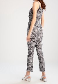Combinaison noire et blanche avec un motif de feuilles ; design sans manches, taille ajustée, jambes fuselées, associée à des chaussures à talons blocs métalliques.