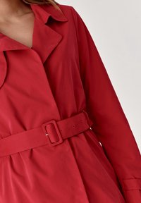 Abrigo rojo con cinturón, hecho de una tela ligera. Presenta un cuello clásico y un diseño de peplum, con una textura suave y una hebilla pulida.