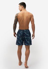 Marineblauwe zwembroek met een tropisch bladenpatroon in teal en koraal. Voorzien van een elastische tailleband en zijzakken.