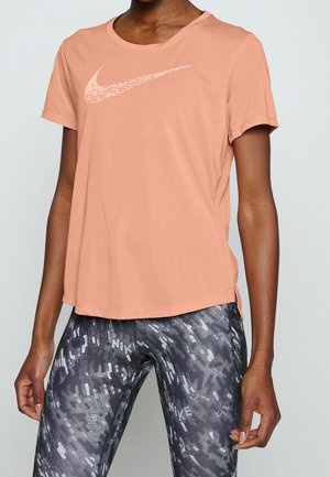 Osoba w brzoskwiniowym t-shircie Nike i czarnych wzorzystych legginsach Nike, stojąca na tle jednolitego jasnego tła. Widoczna górna część ciała.