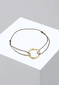 Goldfarbener Metallarmreif mit einem strukturierten, runden Anhänger an einem verstellbaren braunen Band, das ein minimalistisches Design zeigt.