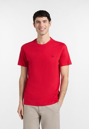 Mladý muž sa usmieva, nosí jednoduché červené tričko s krátkym rukávom a svetlobéžové nohavice, stojí s jednou rukou v vrecku na bielom pozadí.