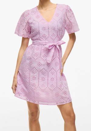 Fioletowa sukienka z ażurowej koronki z krótkimi bufkami, dekoltem w kształcie litery V i kokardą w talii, noszona przez stojącą osobę z jedną ręką na biodrze.