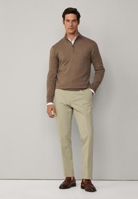 Smeđa zip-up jakna preko bijele košulje s ovratnikom, u kombinaciji sa svijetlosmešavim hlačama i tamnosmešavim loafers cipelama. Jednostavan, prilagođen dizajn.