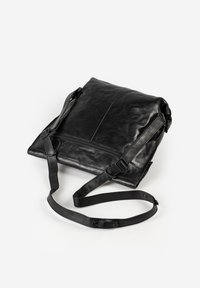 Musta nahkainen crossbody-laukku säädettävällä hihnalla ja etutaskulla, jossa vetoketju, valkoisella taustalla.