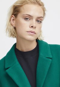 Green wool-blend coat with a notched collar, textured fabric, and black crew neck top underneath. Model has short blonde hair and silver earrings.