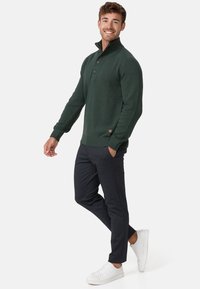 Pull en tricot vert foncé avec un col montant, trois boutons et des poignets côtelés, associé à un pantalon foncé et des baskets blanches.