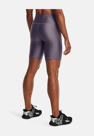 Homme portant un short de sport violet ajusté et taille haute ainsi que des baskets noires Under Armour, debout dos au spectateur.