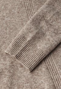 Il dettaglio del maglione lavorato a maglia color marrone chiaro presenta un polsino a coste e un corpo testurizzato, mettendo in risalto un motivo di tessuto morbido e leggermente variegato.