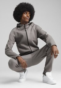 Grå træningsdragt med zip-up hættejakke og bukser, der har en subtil tekstur. Kombineret med hvide sneakers, der fremhæver et minimalistisk design.