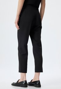 Pantalons noirs ajustés avec une coupe slim, présentant une texture lisse et une finition au niveau de la cheville. Associé à des mocassins en cuir noir.