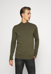 Suéter de cuello alto color caqui hecho de un material suave con textura acanalada, diseño ajustado y mangas largas. Se combina con jeans oscuros.