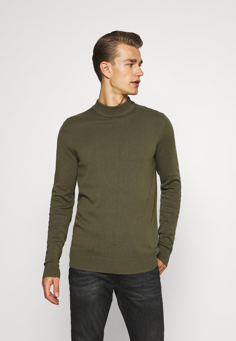 Suéter de cuello alto color caqui hecho de un material suave con textura acanalada, diseño ajustado y mangas largas. Se combina con jeans oscuros.