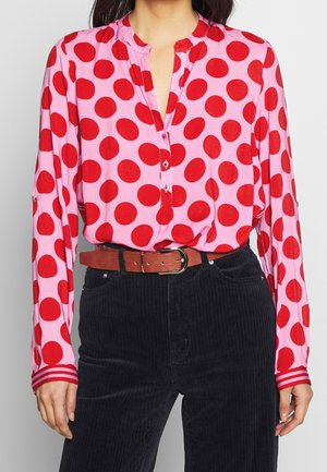 Mujer con blusa de manga larga rosa con grandes lunares rojos, combinada con pantalones de pana negros de talle alto y un cinturón marrón.