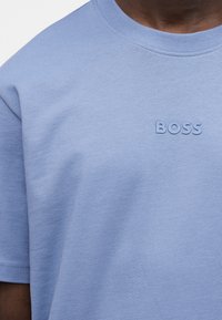 Κοντινό πλάνο ενός ατόμου που φοράει ένα φωτεινό μπλε T-shirt με τη λέξη «BOSS» τυπωμένη σε αντίστοιχα μπλε γράμματα στο στήθος.