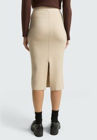 Beżowa spódnica midi wykonana z miękkiego materiału, z tylnym rozporkiem, dopasowanym krojem i gładką teksturą. Noszona z czarnymi skarpetkami i butami.