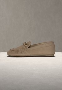 Mocassin en daim beige avec un bout arrondi, des détails cousus autour de la tige et un petit nœud sur le dessus, présenté sur une surface claire.