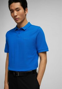 Polo bleu avec un tissu texturé, col à deux boutons et manches courtes. Présente un discret logo brodé sur la poitrine. Porté avec un pantalon noir.