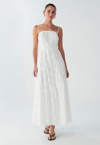 Robe maxi blanche avec un design en couches, tissu œillet texturé, fines bretelles et corsage ajusté. Portée avec des tongs noires.